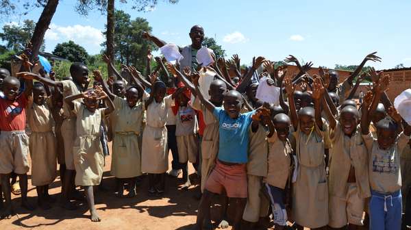 Hilfe für Kinder in Burundi. Bild: Eine Gruppe jubelnder Kinder stehen mit ihrem Lehrer vor einer Schule.