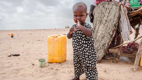 Spenden für Somalia. Bildbeschreibung: Ein Kleinkind steht in einer Wüstenlandschaft, im Hintergrund sind ein Wassercontainer und eine Behausung zu sehen.