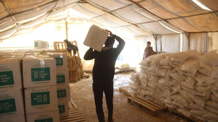 Im Innern eines Zeltes: Links stehen aufgereihte und gestapelte Pakete der Welthungerhilfe. In der Mitte ein Mann von hinten, der ein Paket auf seinen Schultern trägt.