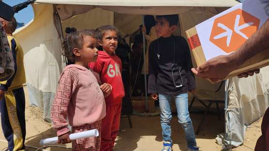 Kinder vor einem Zelt warten auf die Verteilung von Hilfsgütern, von rechts aus dem Bild kommt eine Person mit einer Kiste