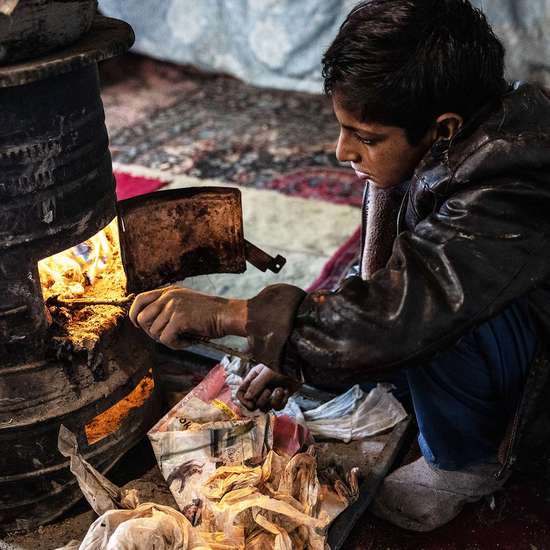 Ein Junge sitzt an einem Ofen und stochert mit einem Ast im Feuer.