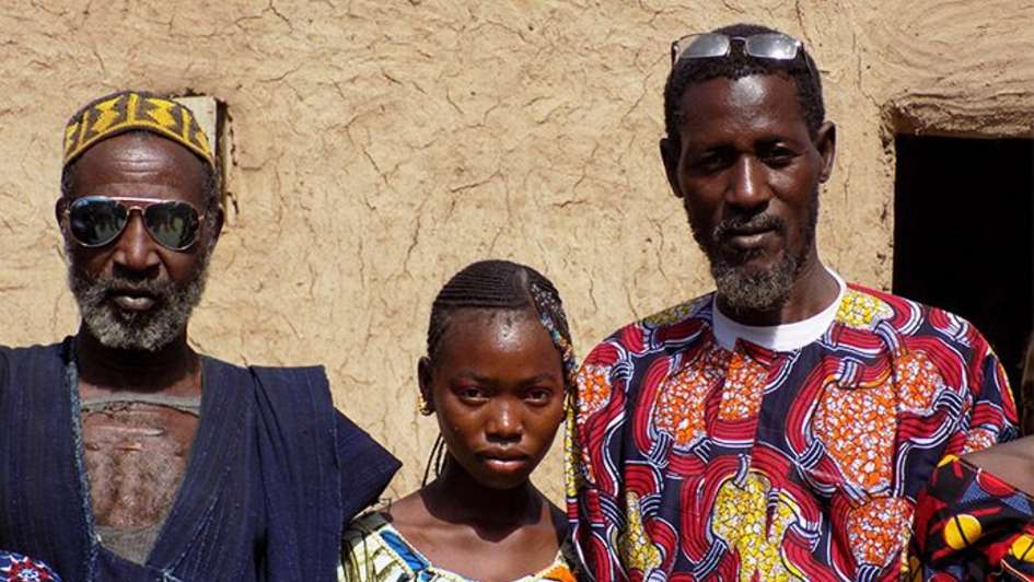 Massiga Diawara vor einer Hausmauer in seinem Dorf einem Mädchen und einem älteren Mann © Schwenk