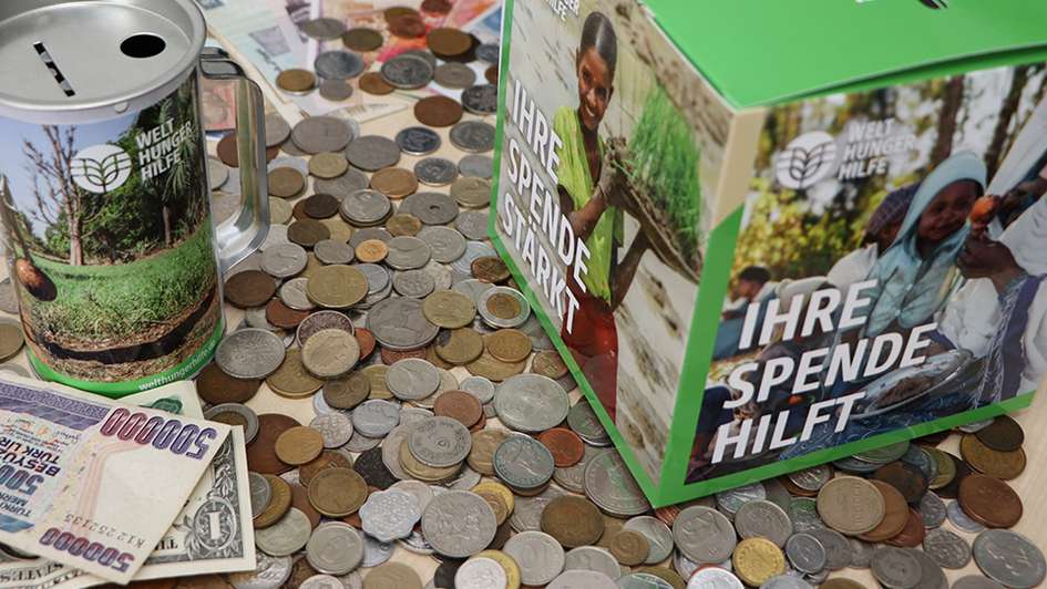 Symbolbild: Spendenbox und Spendendose der Welthungerhilfe, umgeben von Münzen und Scheinen unterschiedlicher Währungen