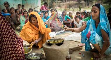 Zwei Frauen kochen, im Hintergrund schauen mehrere Frauen zu.