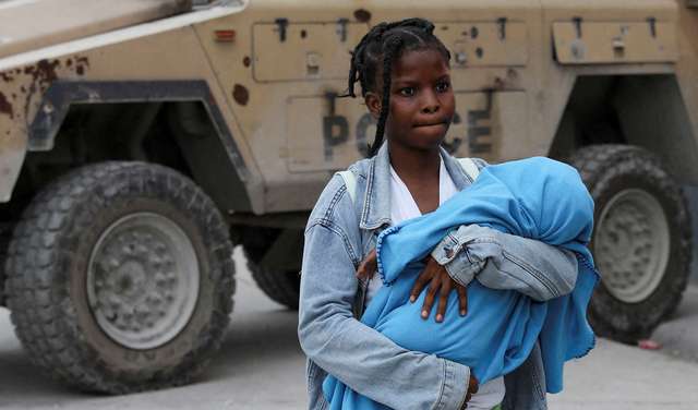 Eine junge Frau in Port-au-Prince, Haiti, verlässt mit ihrem Baby im Arm ihr Zuhause auf der Flucht vor Bandengewalt, im Hintergrund ein gepanzertes Fahrzeug