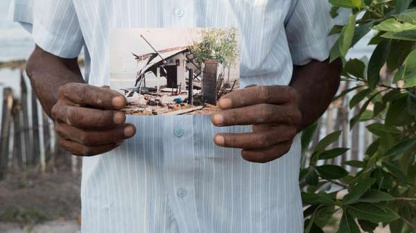 Nahaufname eines Mannes in weißer Kleidung, der ein Foto seines zerstörten Hauses in der Hand hält.