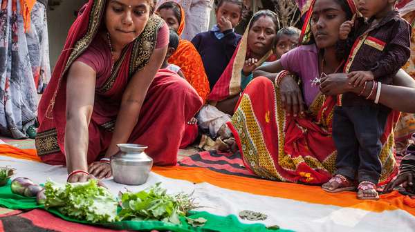 Eine Frau in einem Sari hockt auf dem Boden, vor ihr eine indische Flagge. Sie schiebt einen Salat auf den grünen Teil der Flagge.