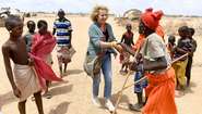 Eine Person aus Marsabit, Kenia, schüttelt Michaela May die Hand. Sie sind umgegen von Kindern und einem jungen Mann aus der Region. Im Hintergrund ist eine trockene Landschaft zu sehen.