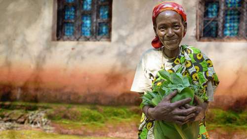 Eine ältere Frau hält Maniokblätter in ihren Armen und lacht in die Kamera.