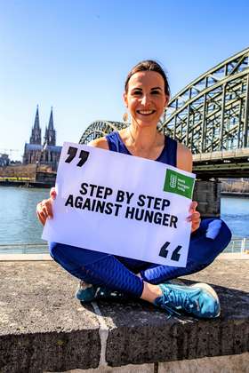 Sabrina Mockenhaupt-Gregor sitzt auf einer Mauer und hält ein Schild zum #ZeroHungerRun in der Hand. Im Hintergrund sieht man den Kölner Dom.