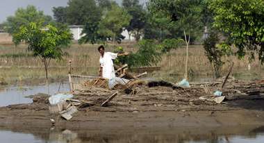 Ein Mann steht auf einem von Wasser umgebenen Stück Land und zeigt auf zerstörte Haushaltsgegenstände.
