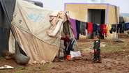 Zwei Kinder stehen vor einem Zelt in einem Flüchtlingscamp und schauen Richtung Kamera. An einer Leine ist Wäsche aufgehängt.