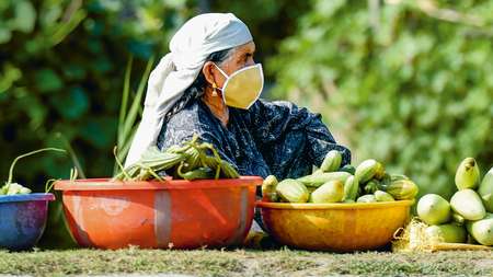 Eine Frau mit Mundschutz und einem Tuch um den Kopf sitzt auf dem Boden, neben sich stehen Schüsseln mit Gemüse.