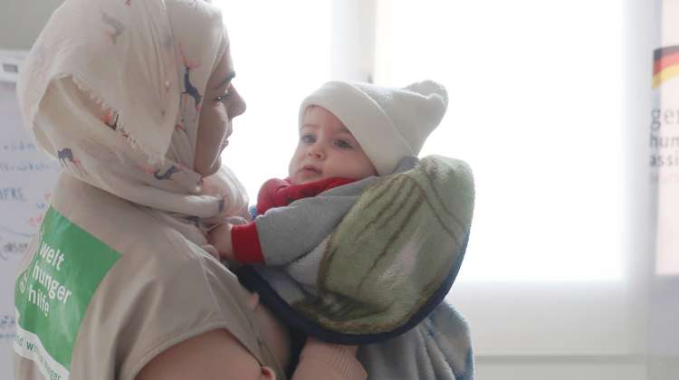 Eine Frau mit Kopftuch und Welthungerhilfe-Weste hält ein Kleinkind auf dem Arm