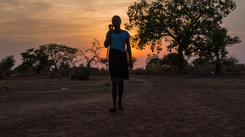 Hilfe für den Südsudan. Bildbeschreibung: Eine Frau geht auf die Kamera zu, im Hintergrund sieht man den Sonnenuntergang.