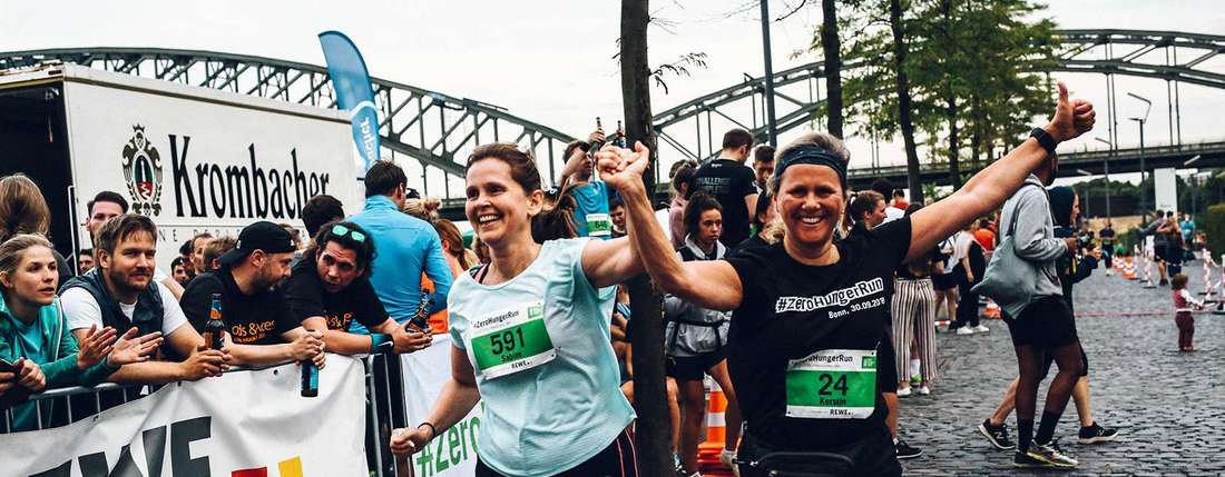 Zwei laufende Frauen in Sportkleidung heben jubelnd ihre Arme in die Höhe. Im Hintergrund sind Zuschauer und der Kölner Rheinauhafen zu sehen. 