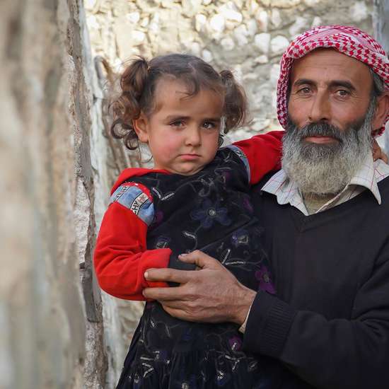 Ein älterer Mann mit einem kleinen Mädchen auf dem Arm. Links oben steht in einem Logo: "Syrien. 10 Jahre Krieg"