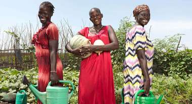 Drei Frauen in einem landwirtschaftlichen Garten halten Gießkannen und lächeln in die Kamera.