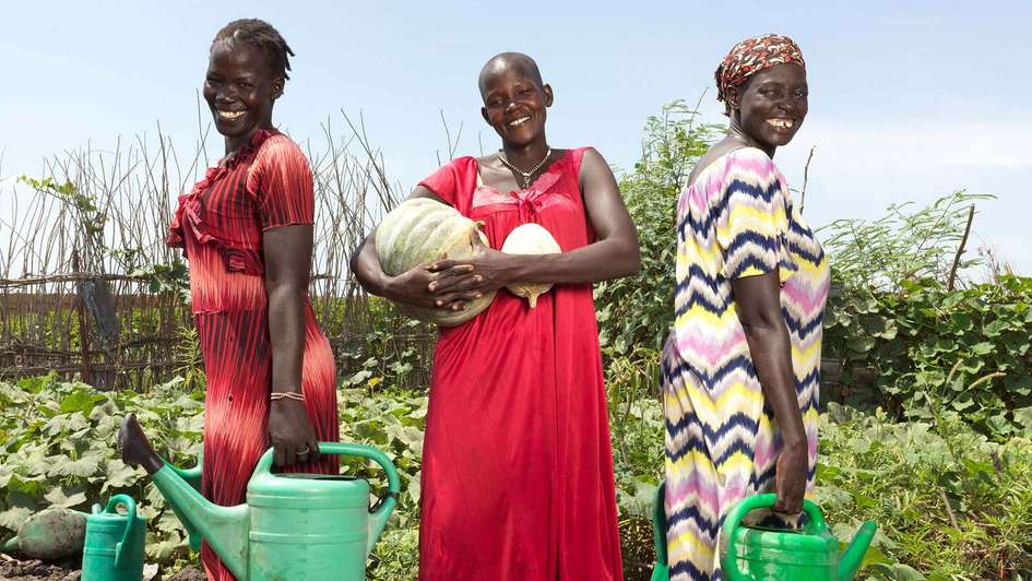 Drei Frauen in einem landwirtschaftlichen Garten halten Gießkannen und lächeln in die Kamera.