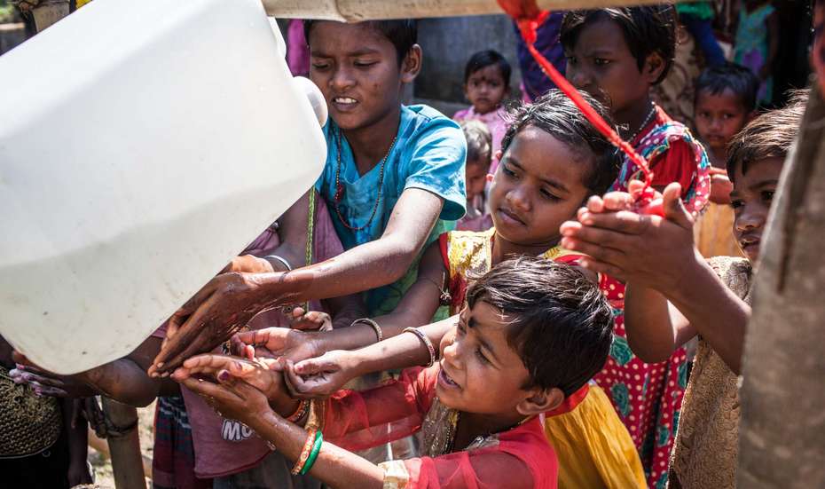Aus einem Kanister wird Wasser geschütet, damit sich die Kinder ihre Hände vor dem Essen waschen können, Indien.
