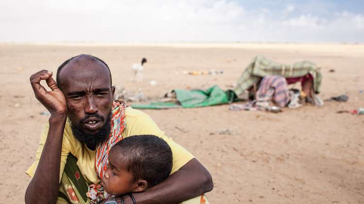 Ihre Hilfe für Somalia. Bildbeschreibung: Ein erschöpft aussehender Mann und ein Kleinkind hocken in einer Wüstenlandschaft. 