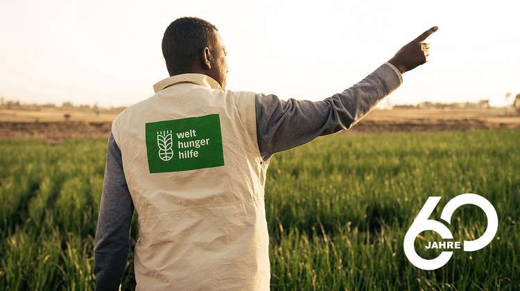 Ein Mitarbeiter ist von hinten zu sehen, mit der rechten Hand zeigt er nach rechts oben. Er trägt eine beige Weste mit grünem Welthungerhilfe-Logo.