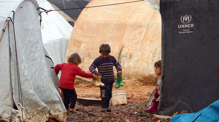 Drei Kinder stehen zwischen Zelten in einem Flüchtlingscamp. Sie halten Gummistiefel in ihren Händen.