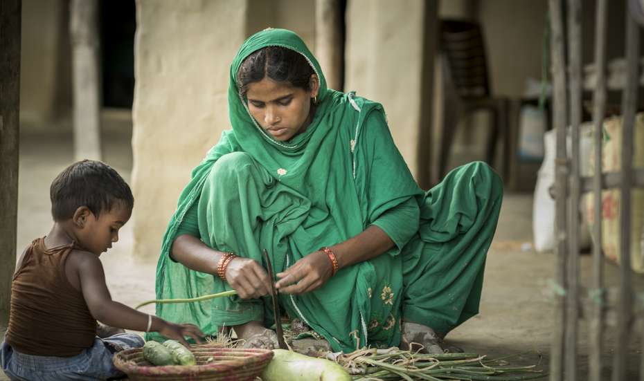 Eine Frau hockt auf dem Boden und bearbeitet Gemüse, neben ihr sitz ein Kind.