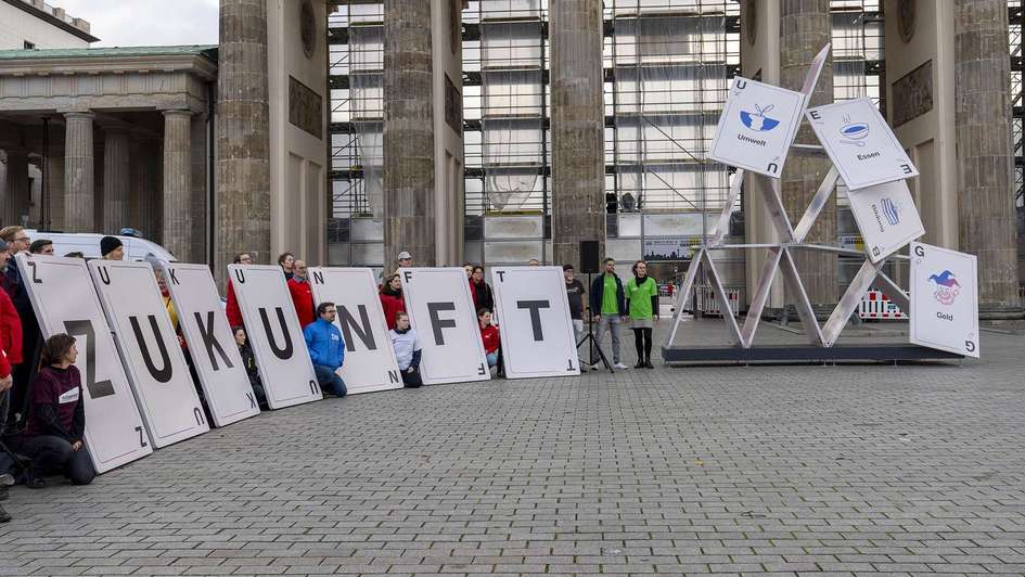 Aktionsgruppe vor dem Brandenburger Tor vor einem symbolisch einstürzenden Kartenhaus. Sie halten große Karten mit der Aufschrift "Zukunft".