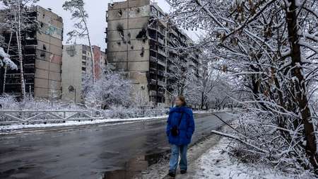 Eine Frau läuft eine Straße entlang, im Hintergrund zerstörte Wohnhäuser, es ist kalt und verschneit