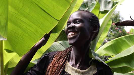 Bild aus einem Projekt im Südsudan mit Schulmahlzeiten, eine Frau steht unter einer Palme und lächelt