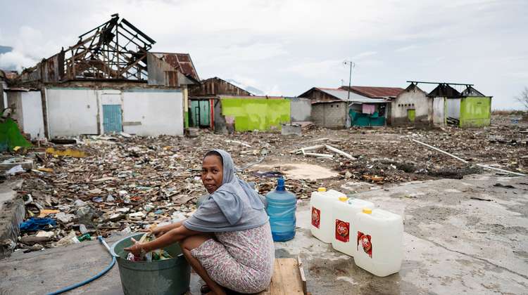 Eine Frau sitht auf einem Stück Holz, vor ihr ein Wäschekübel. Um sie herum sieht man die Zerstörung, die der Tsunami angerichtet hat.