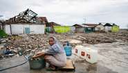 Eine Frau sitht auf einem Stück Holz, vor ihr ein Wäschekübel. Um sie herum sieht man die Zerstörung, die der Tsunami angerichtet hat.