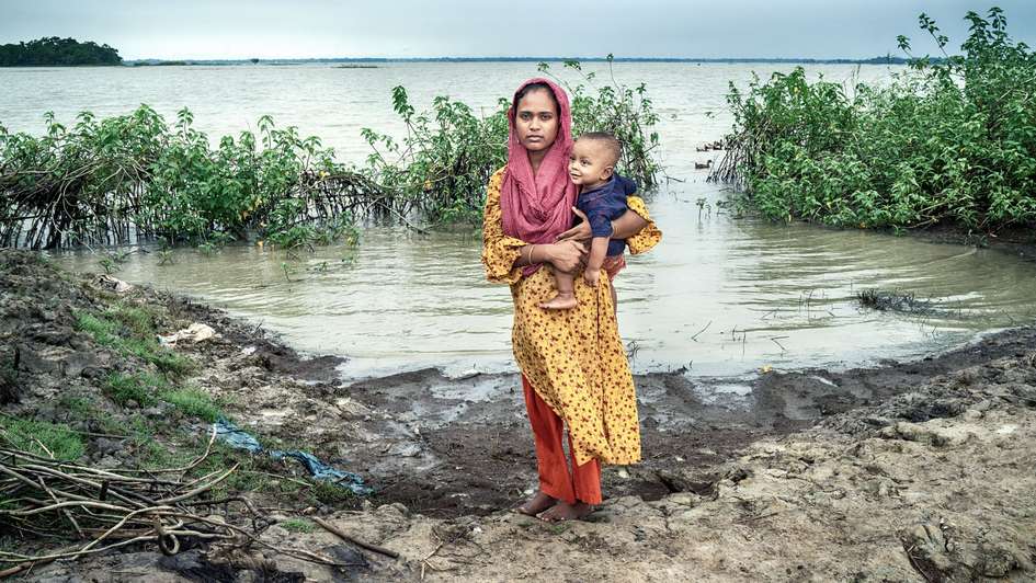 Eine Mutter mit ihrem Kind auf dem Arm steht vor überschwemmtem Land in Bangladesch.