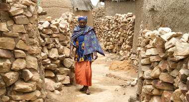 Eine Frau in Mali läuft zwischen Lehmhütten entlang.