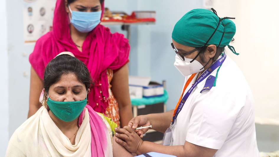 Es sind drei indische Frauen abgebildet: Eine Ärztin gibt einer Frau eine Spritze, eine weitere Frau schaut zu. Sie alle tragen Masken.