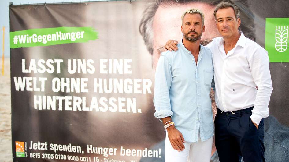 Die Schauspieler Stephan Luca und Jan Sosniok vor einem Plakat der Welthungerhilfe. Auf dem Plakat steht "Lasst uns eine Welt ohne Hunger hinterlassen".