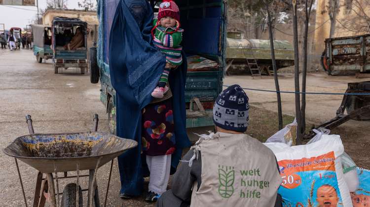 Eine Frau mit blauer Burka steht vor einem Welthungerhilfe Mitarbeiter. Sie hält ein kleines Kind im Arm. 
