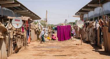 Straße in einem Flüchtlingscamp im Sudan, links und rechts reihen sich Unterkünfte, in der Mitte spannt eine Wäscheleine mit einem großen purpurnem Tuch.