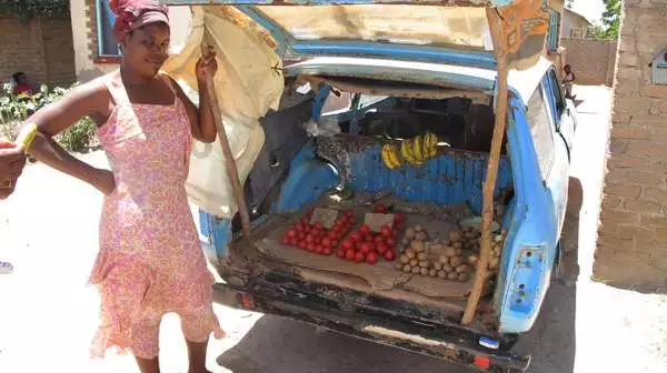 Marktfrau vor ihrem, mit Obst und Gemüse beladenen, Kofferraum.