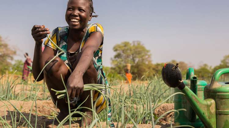 Ihre Hilfe für den Südsudan - jetzt spenden. Bild: Eine Frau kniet lachend auf einem Gemüsebeet und hält eine Pflanze in der Hand.