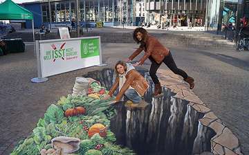 Illusionsmalerei als Straßenkunst: Zwei Frauen balancieren über eine Schlucht zu Gemüse und Obst.