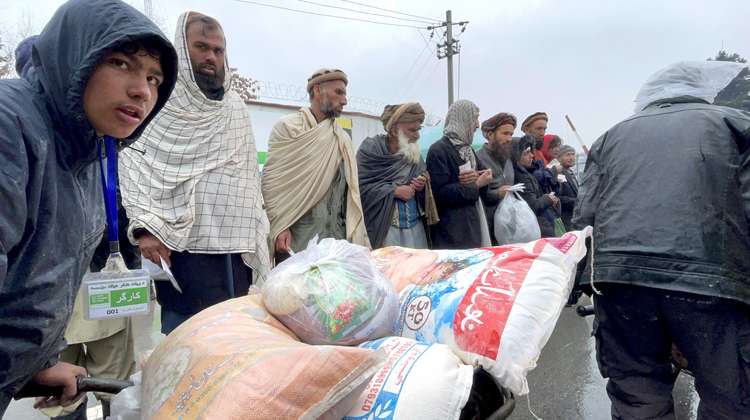 Männer stehen bei einer Lebensmittelverteilung in Afghanistan nebeneinander, während ein junger Mann eine Schubkarre mit Säcken vorbei schiebt.