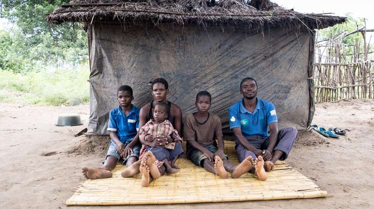 Ein Mann, eine Frau und drei Kinder sitzen auf einer Bastmatte, hinter ihnen eine Hütte aus Planen.