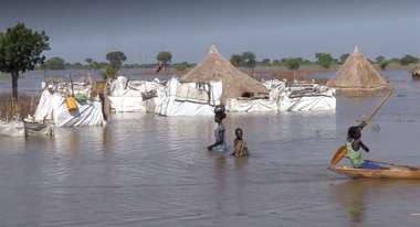 Überschwemmungen im Südsudan: Eine Frau und ein Kind waten durch das hüfthohe Wasser, im Hintergrund sind überschwemmte Hütten zu sehen.