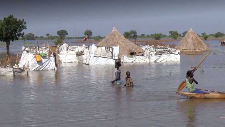 Überschwemmungen im Südsudan: Eine Frau und ein Kind waten durch das hüfthohe Wasser, im Hintergrund sind überschwemmte Hütten zu sehen.