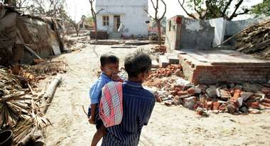 Ein Mann von hinten, er trägt ein Kind auf dem Arm, das Kind schaut in die Kamera. Im Hintergrund sind zerstörte Häuser.