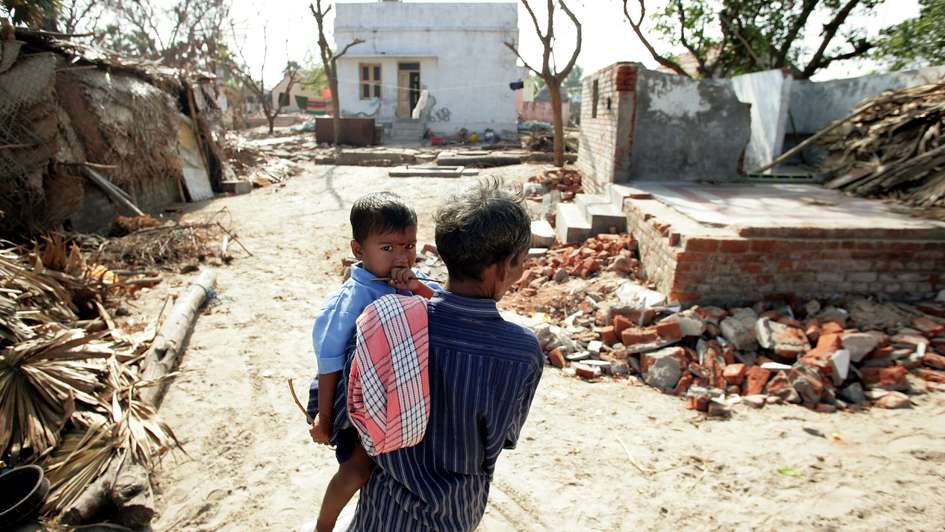 Ein Mann von hinten, er trägt ein Kind auf dem Arm, das Kind schaut in die Kamera. Im Hintergrund sind zerstörte Häuser.