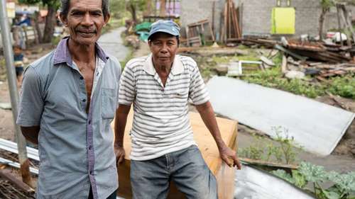 Ihre Hilfe für Indonesien. Bildbeschreibung: Zwei Männer schauen in die Kamera, hinter ihnen sieht man zerstörte Häuser.