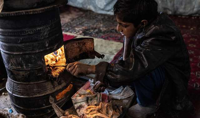 Ein Junge sitzt an einem Ofen und stochert mit einem Ast im Feuer.